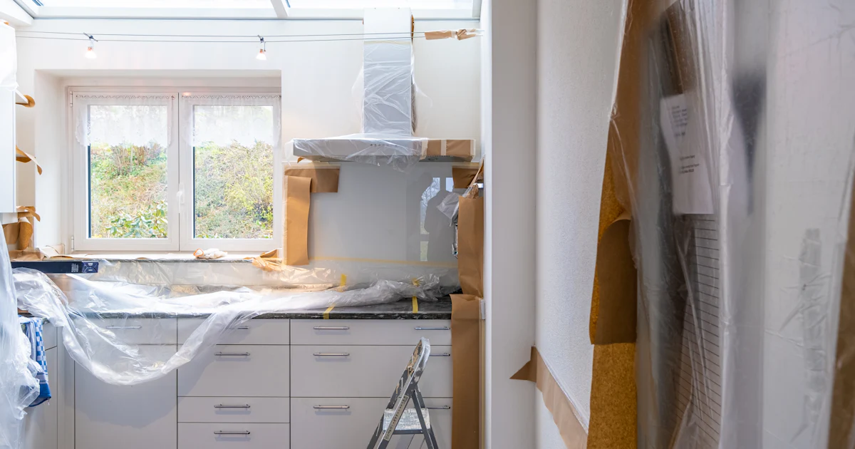 A kitchen in the middle of a renovation, cabinets and counters masked with plastic sheeting and brown paper, a step ladder on the floor