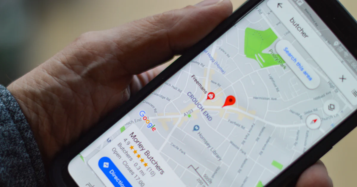 A hand holding a phone showing a Google Maps search for a local butcher, with the top result's star rating, distance and opening hours